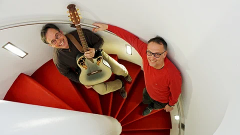 Zwei Männer auf roter Wendeltreppe, einer hält Gitarre.