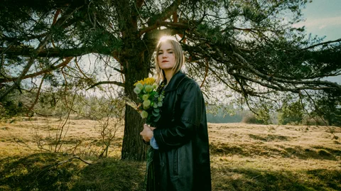 Frau mit Blumenstrauss vor einem Baum auf einer Wiese.