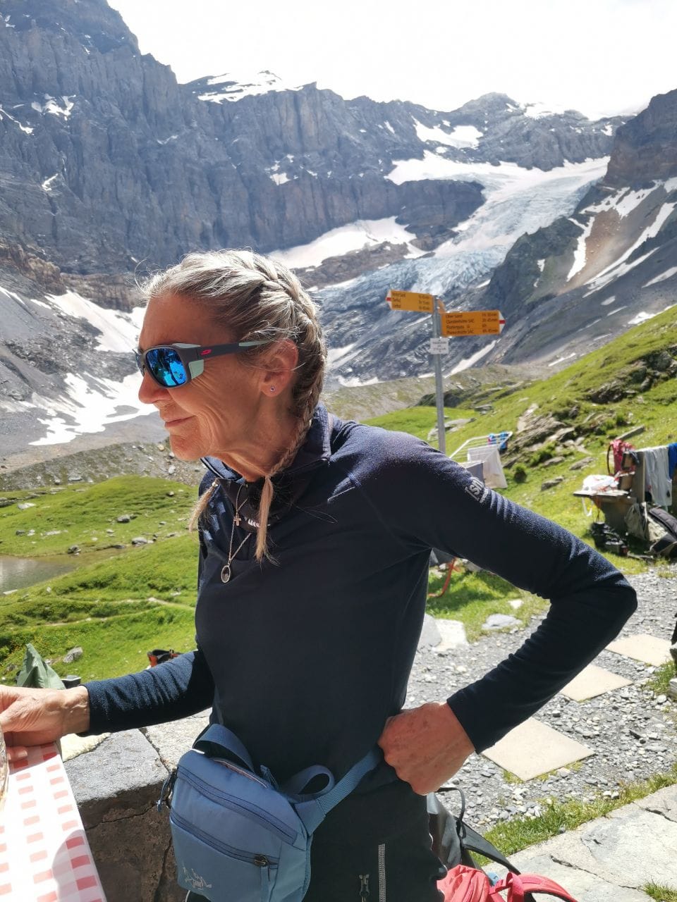 Frau mit Sonnenbrille und Zopf vor Berglandschaft mit Gletscher und Wegweisern.