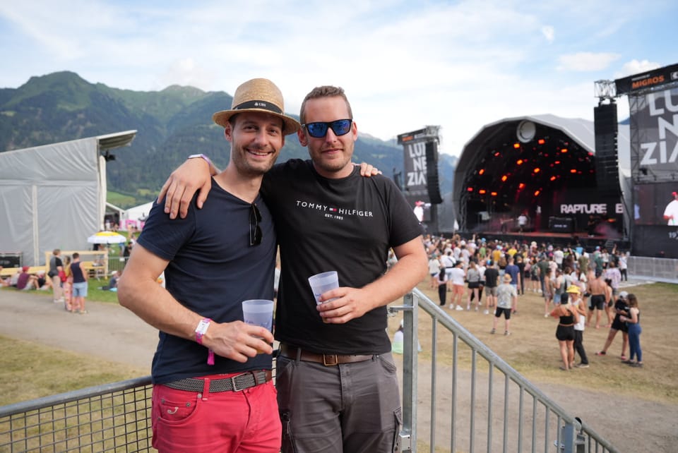 Zwei Männer mit Getränkebechern bei einem Musikfestival vor einer Bühne.