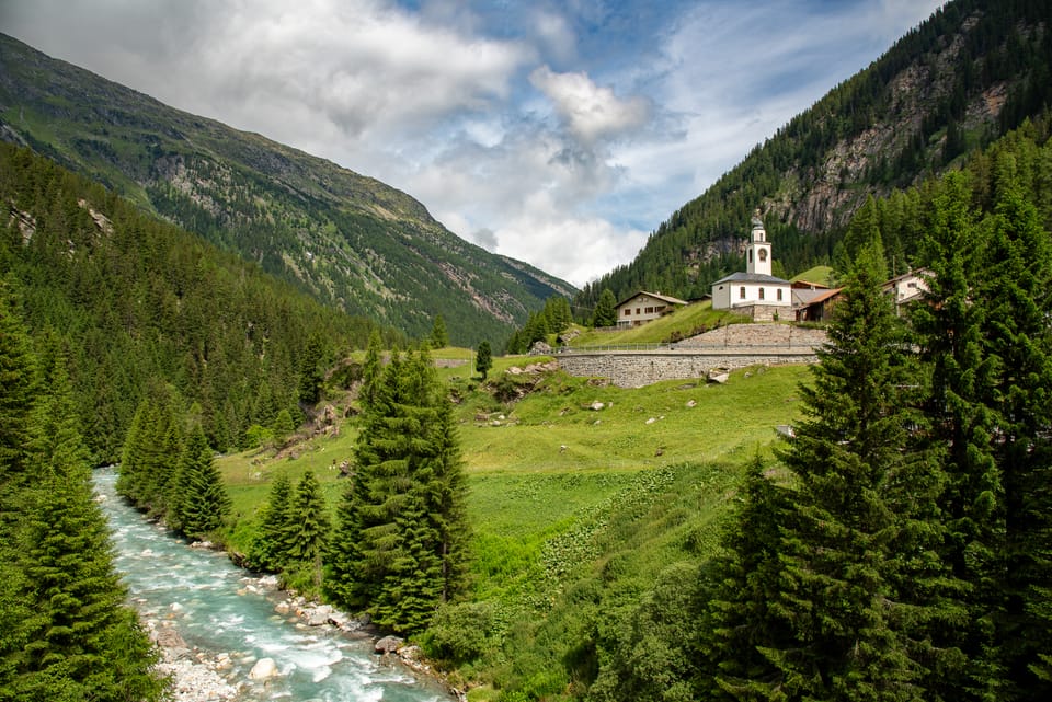 Bach in alpiner Landschaft mit Kirche und Bergen.