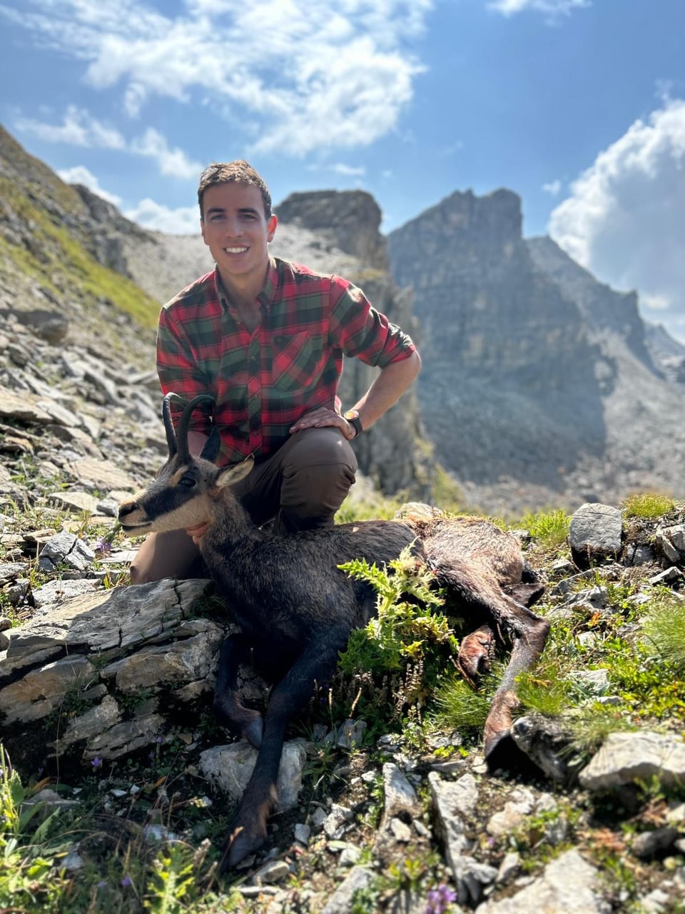 Mann in kariertem Hemd posiert mit einem erlegten Wildtier in bergiger Landschaft.