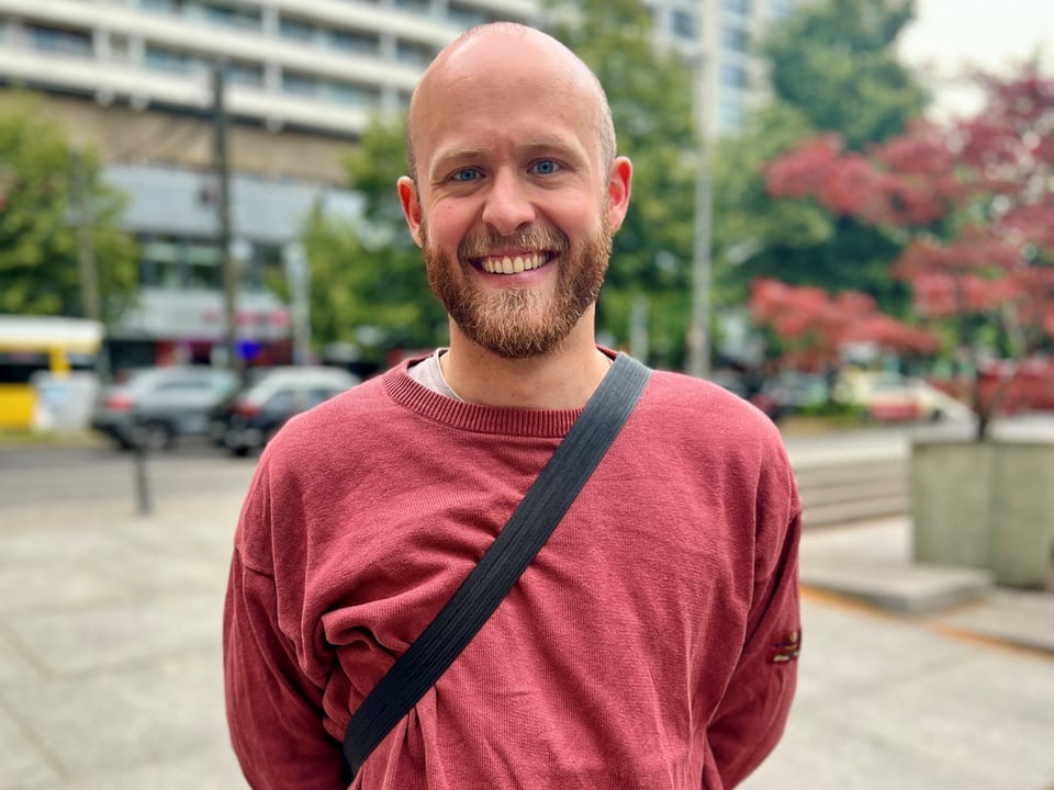 Mann in rotem Pullover lächelnd im Freien vor unscharfem städtischem Hintergrund