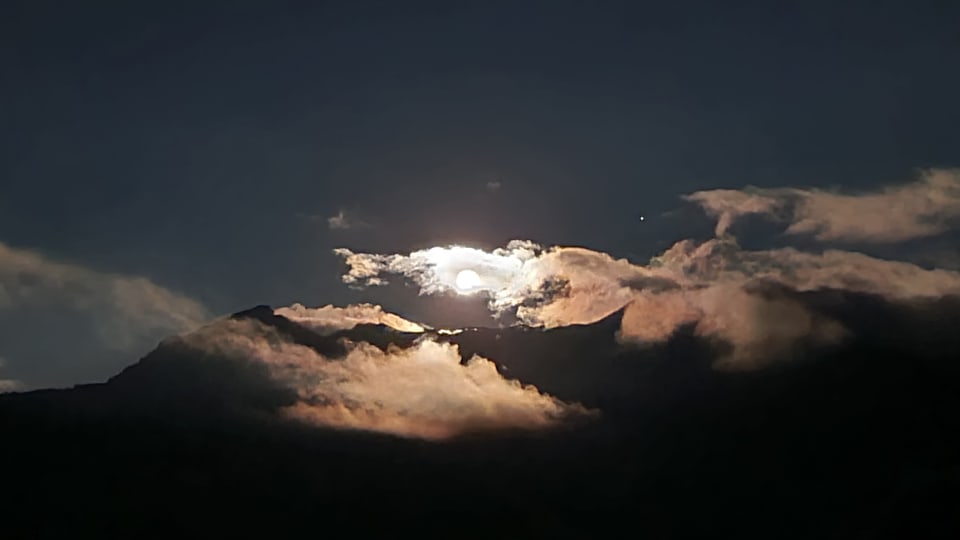 Nachtaufnahme des Mondes, der hinter Wolken und einem Berg hervorschaut.