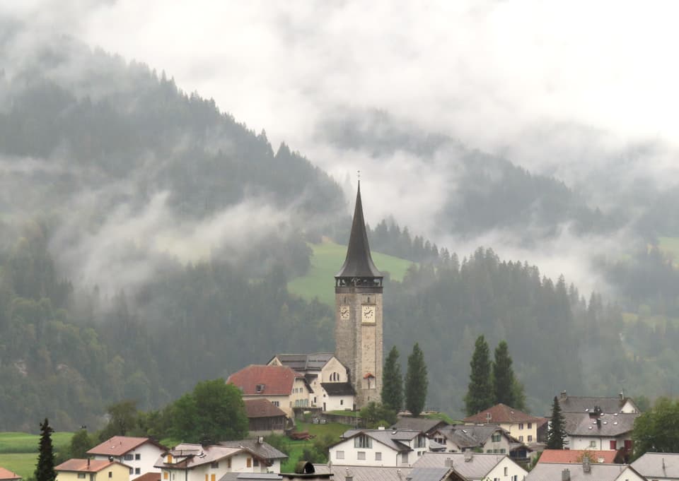 Dorf mit Kirchturm und Bergen im Nebel.