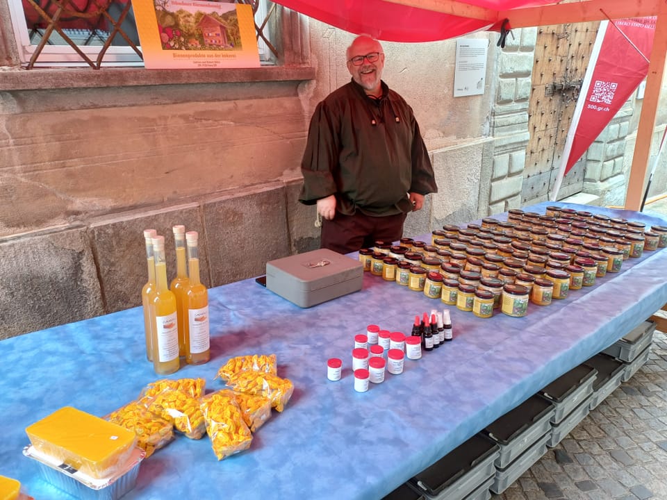 Mann steht hinter einem Marktstand mit verschiedenen Produkten unter einem roten Zelt.
