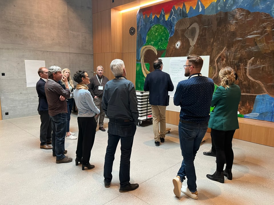 Gruppe von Menschen steht vor einem Whiteboard in einem Raum mit grossem Wandgemälde.