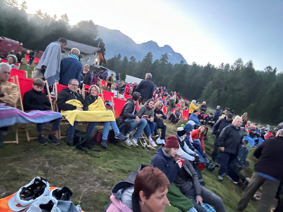 Menschen sitzen auf Stühlen und Gras bei einer Outdoor-Veranstaltung in den Bergen.