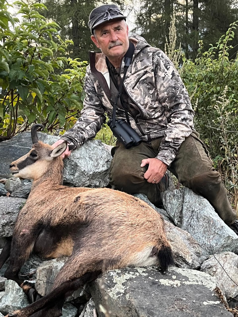 Mann mit Tarnjacke neben stehendem Bock auf Felsen.