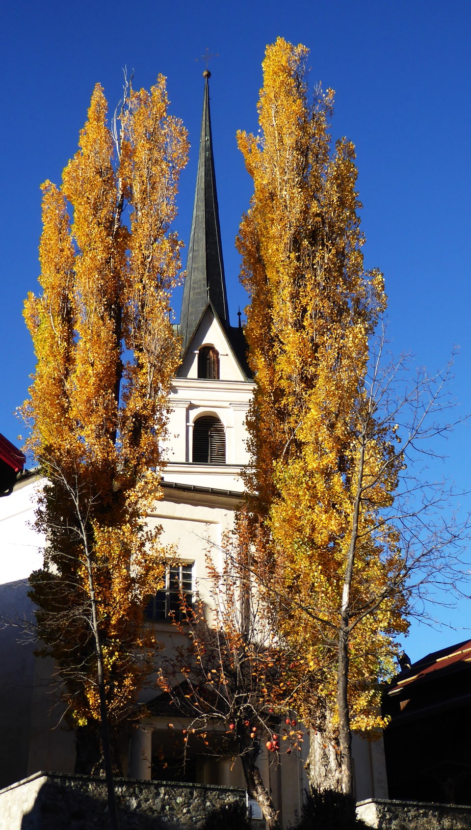 Kirchturm hinter gelben Herbstbäumen vor blauem Himmel.