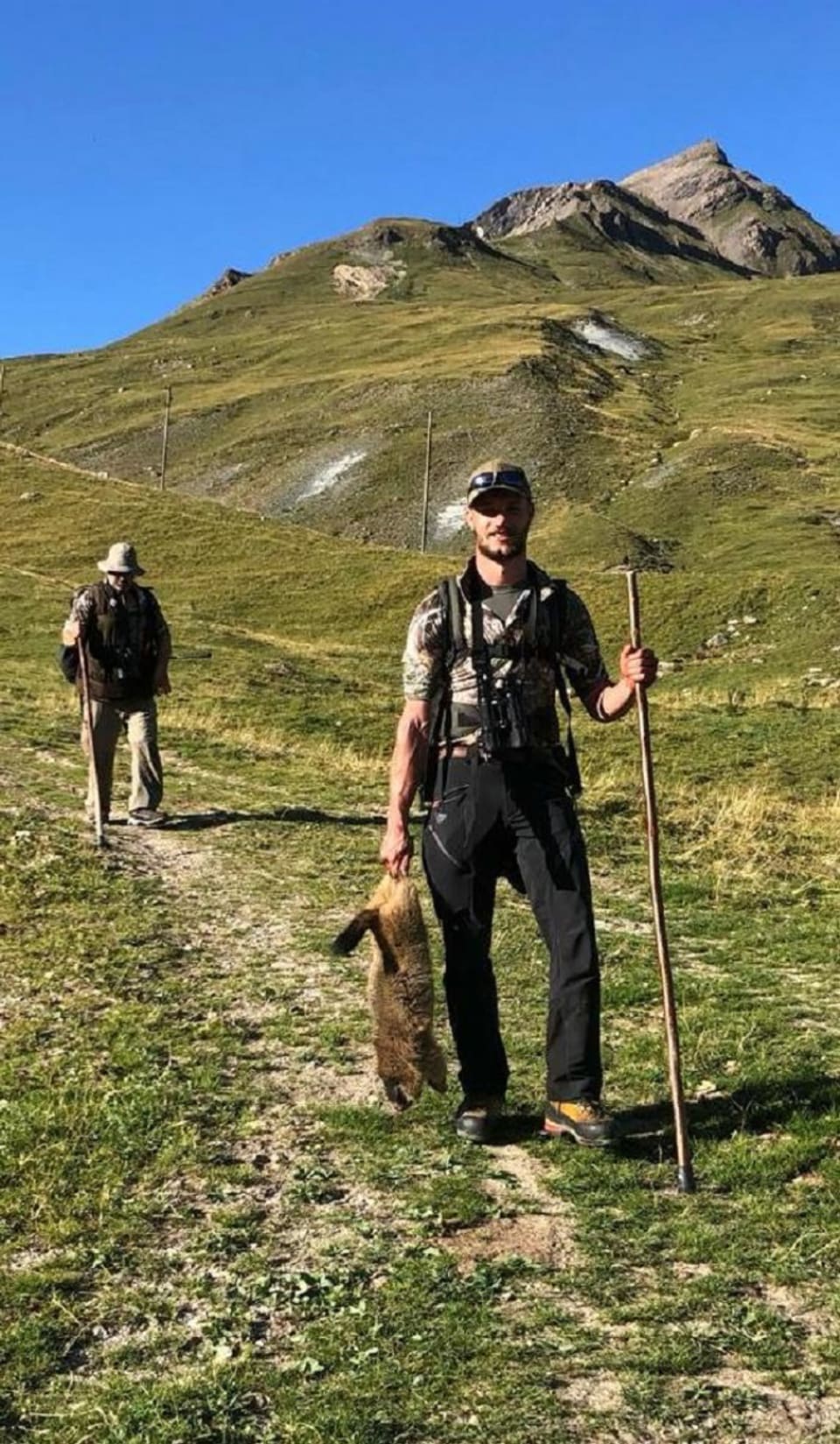 Ein Jäger trägt ein geschossenes Murmeltier, gefolgt von einem Jagdbegleiter.