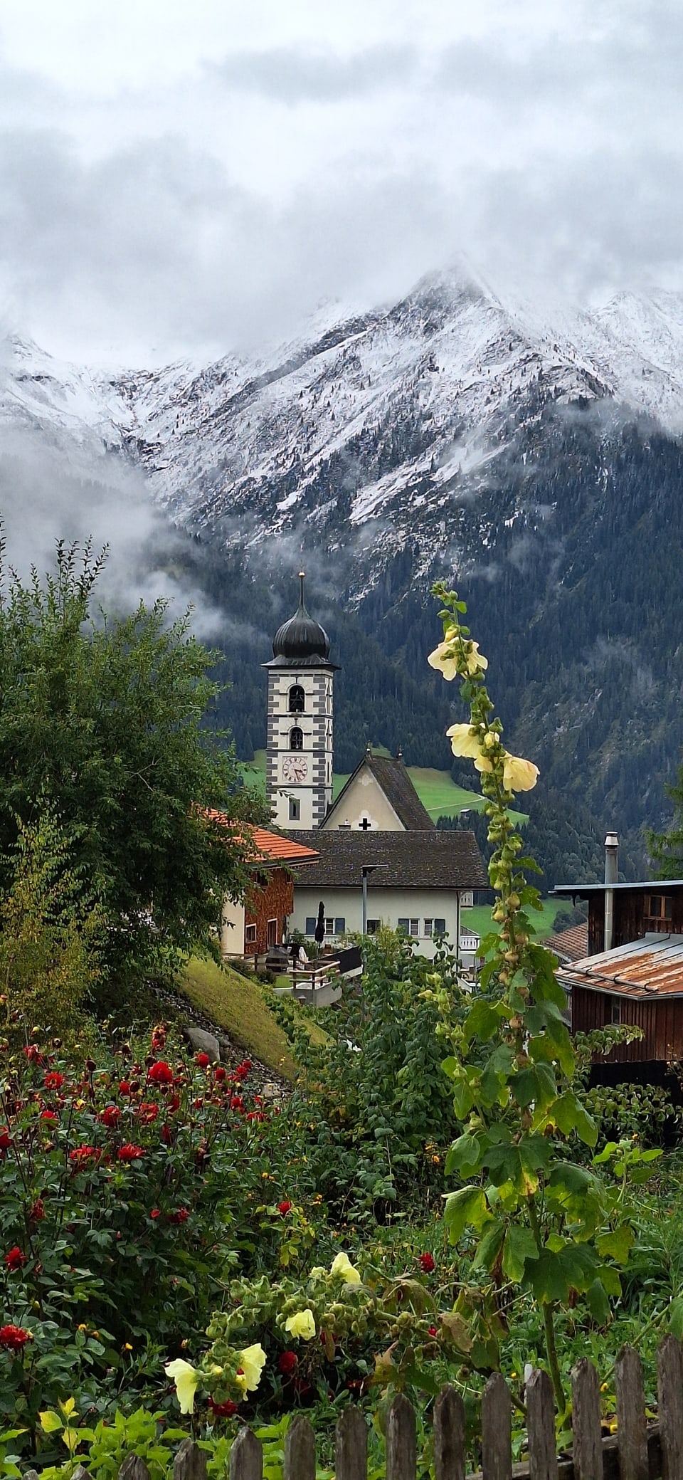 Kirchturm in einem Alpen-Dorf mit schneebedeckten Bergen im Hintergrund.