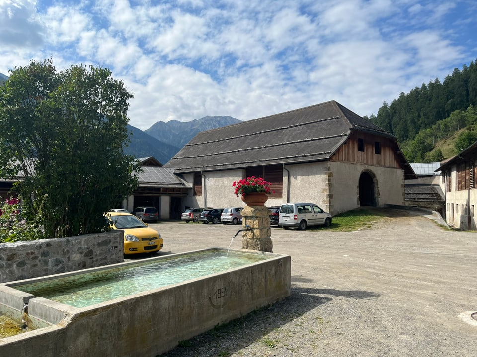 Bauernhof mit Brunnen und Autos vor Bergen.