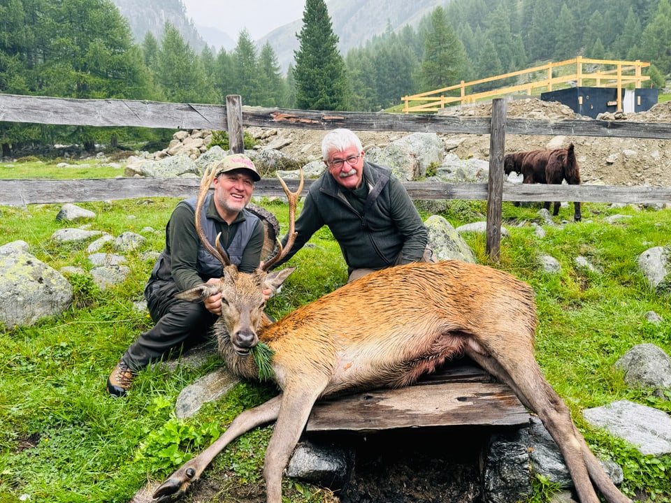 Zwei Männer posieren mit einem erlegten Hirsch auf einer Wiese.