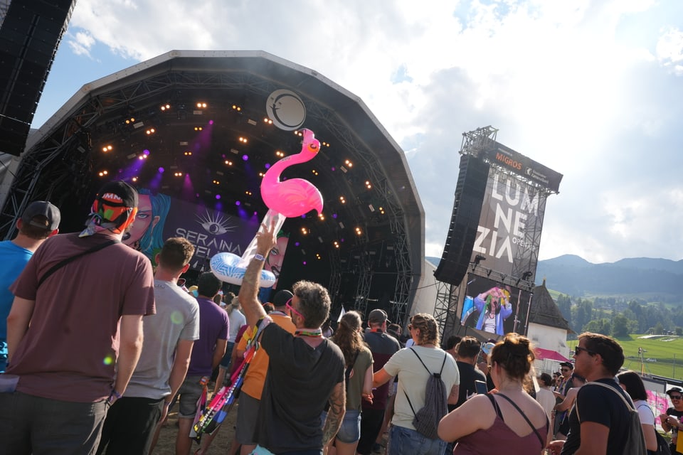 Menschenmenge bei einem Open-Air-Konzert mit Bühne und Bergen im Hintergrund am Open Air Lumnezia 2024.