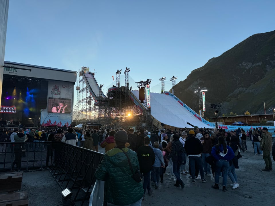 Menschenmenge vor Bühne und Snowboardrampe im Freien bei Abenddämmerung.