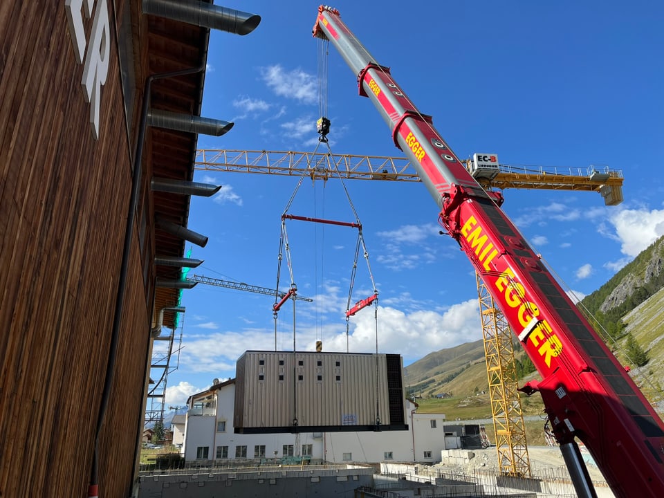 Kran hebt grosses Containerhaus neben Holzwand und Berglandschaft.