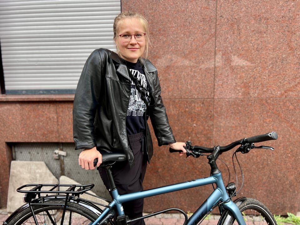 Person mit schwarzer Lederjacke steht neben einem blauen Fahrrad.