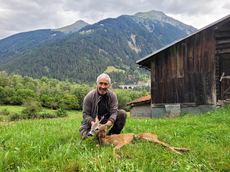 Mann kniet neben einem Reh auf einer Wiese vor einem Holzhaus und Bergen.