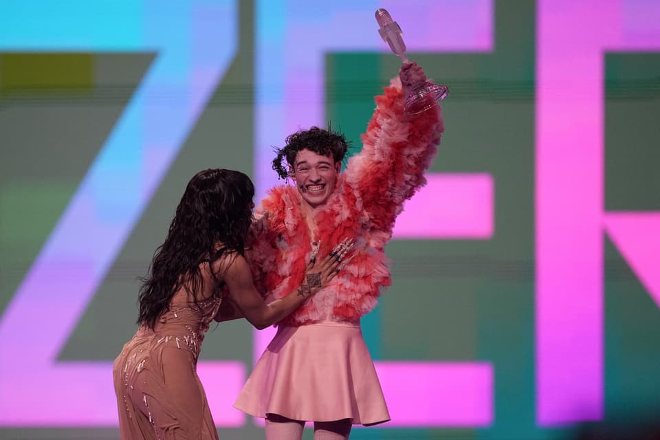 Person in rosa Kleid und orangefarbener Federjacke erhält einen Award von einer Frau auf einer Bühne.