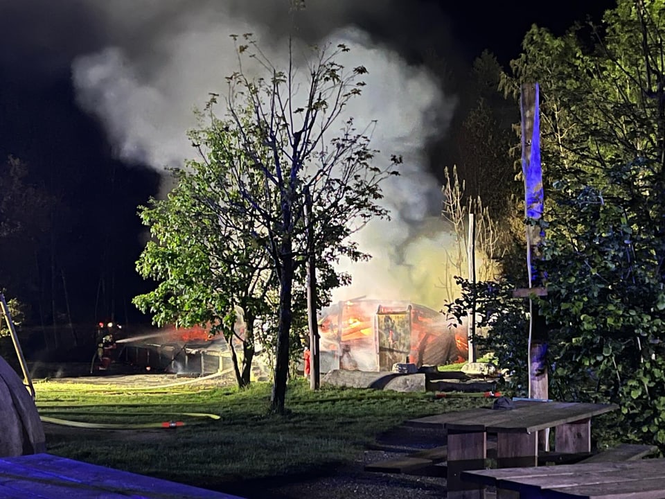 Nächtliche Szene mit brennender Holzhütte und Rauch, umgeben von Bäumen.