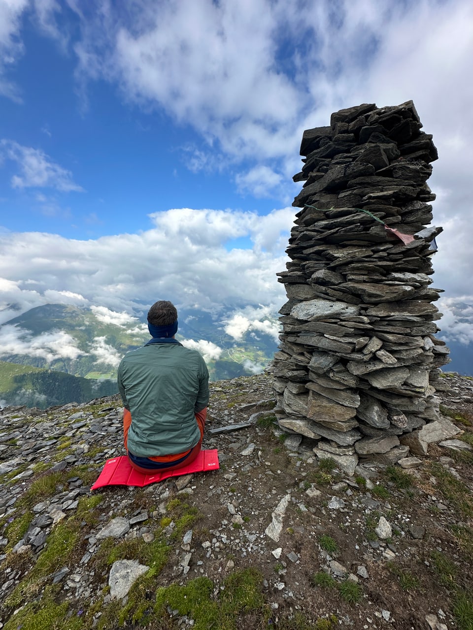 Person sitzt vor Steinhaufen auf Berggipfel mit Wolkenhimmel.