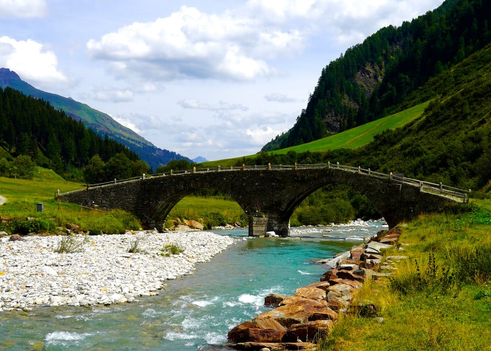 Steinbrücke über einen klaren Fluss in einer Berglandschaft.