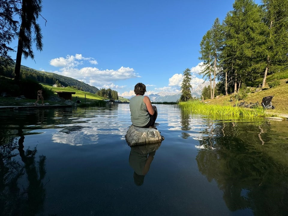 Person sitzt auf einem Felsen im See, umgeben von Bäumen.