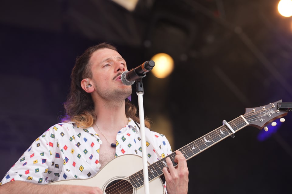 Musiker singt in ein Mikrofon und spielt Gitarre auf der Bühne am Open Air Lumnezia 2024.