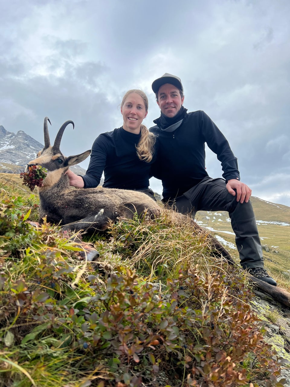 Zwei Personen posieren mit erlegtem Gämsbock in alpiner Landschaft.