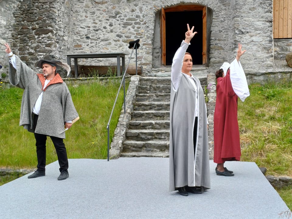Drei Figuren in historischen Kostümen führen ein Schauspiel im Freien vor einer Steinwand auf.