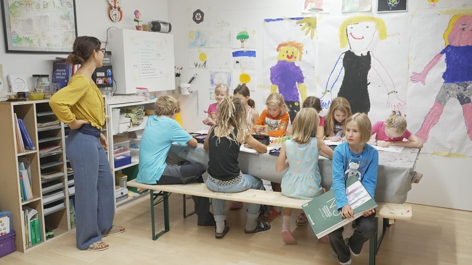 Kinder sitzen an einem langen Tisch, im Hintergrund sind Zeichnungen aufgehängt. 