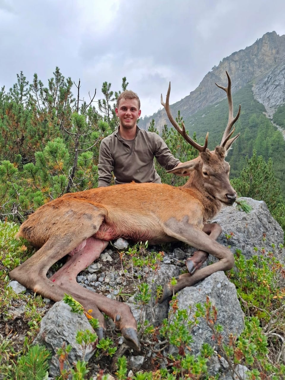 Mann mit erlegtem Hirsch auf einem Felsen in bergiger Landschaft.