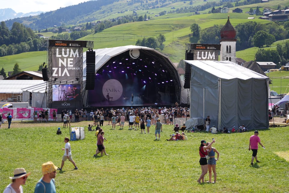 Festivalbesucher vor einer Bühne im Freien mit Bergen im Hintergrund am Open Air Lumnezia 2024 in Degen.