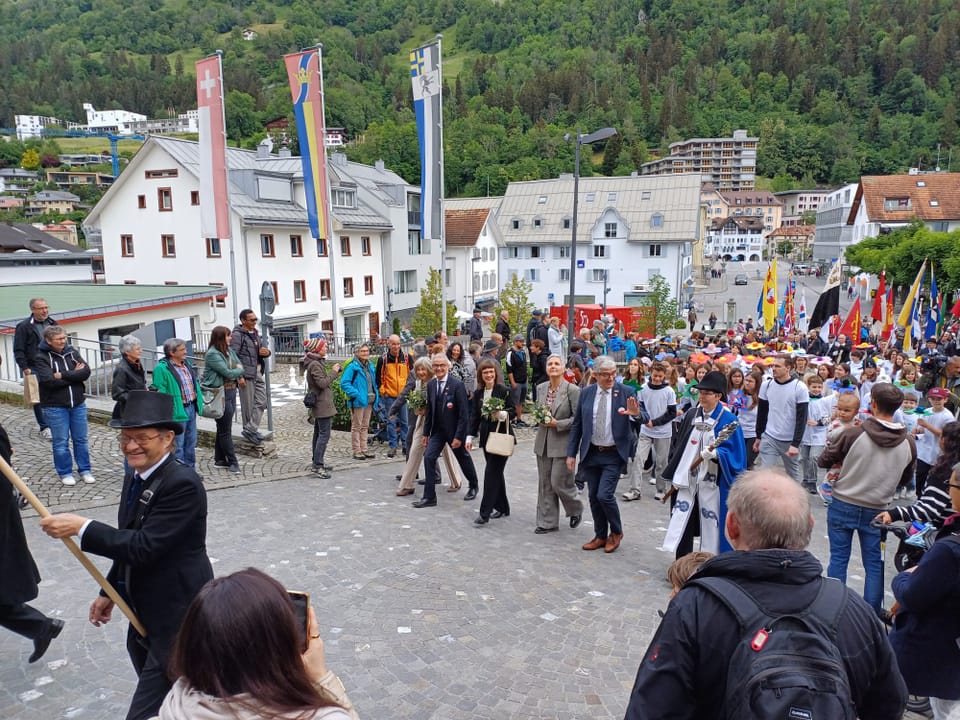 Menschenmenge bei einem Stadtfest vor Häusern und bewaldeten Hügeln.