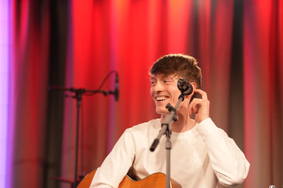 Lächelnder Mann mit Gitarre auf einer Bühne mit roten Vorhängen.