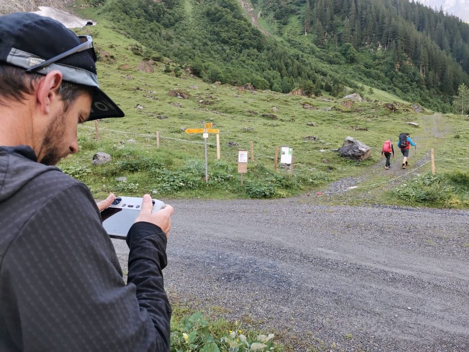 Mann steuert Drohne in bergiger Landschaft; Wanderer im Hintergrund.