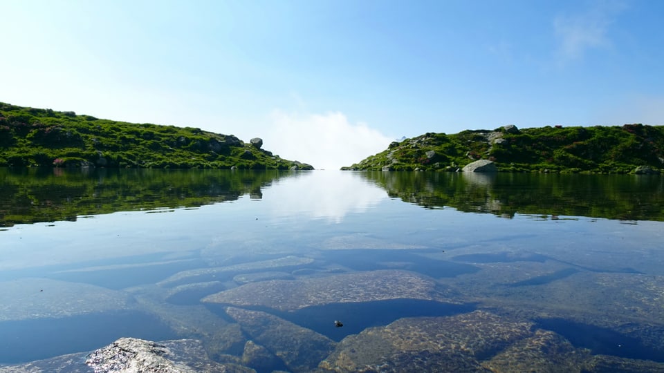 Klares Bergsee-Wasser mit grünen Hügeln im Hintergrund.