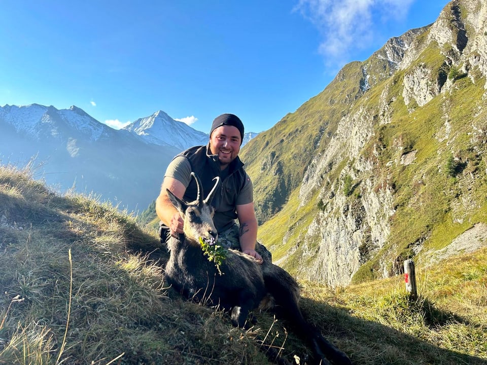 Mann sitzt mit einem Tier in Berglandschaft.