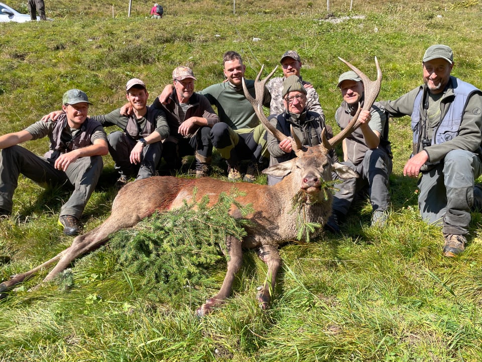 Gruppe von acht Jägern mit einem erlegten Hirsch auf einer Wiese.