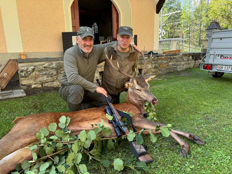 Zwei Männer posieren mit einem erlegten Hirsch vor einem Gebäude.