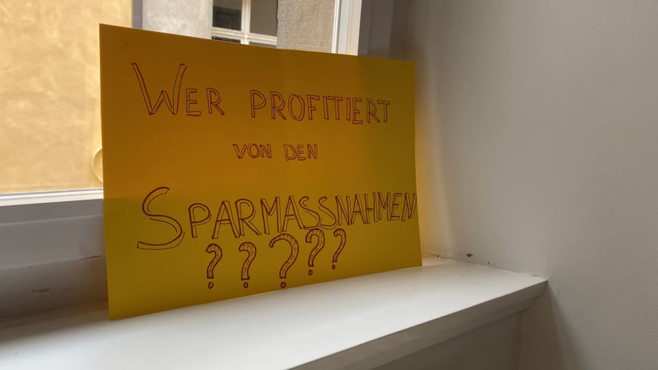 Gelbes Schild mit der Aufschrift 'Wer profitiert von den Sparmassnahmen?????' auf einer Fensterbank.