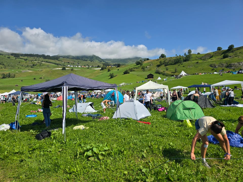 Campingszene auf einer grünen Wiese mit Hügeln und Zelten im Hintergrund.