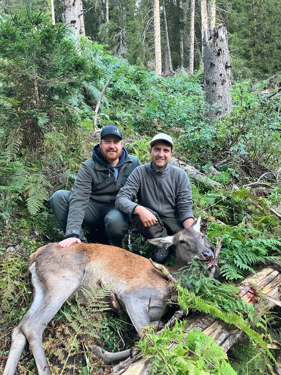 Zwei Männer posieren mit einem erlegten Hirsch im Wald.