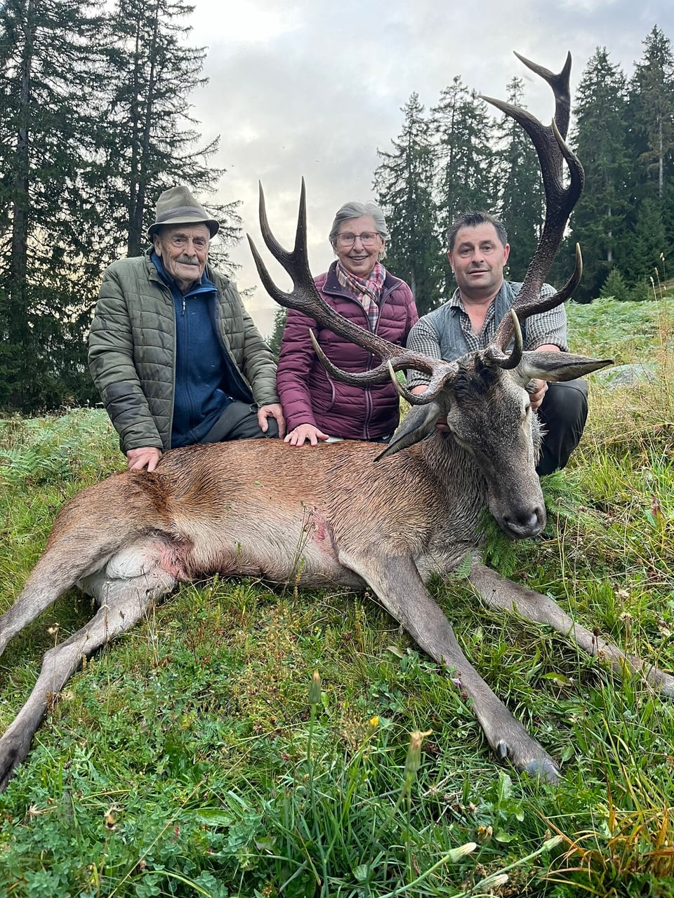 Drei Personen posieren mit einem erlegten Hirsch auf einer Wiese vor einem Wald.