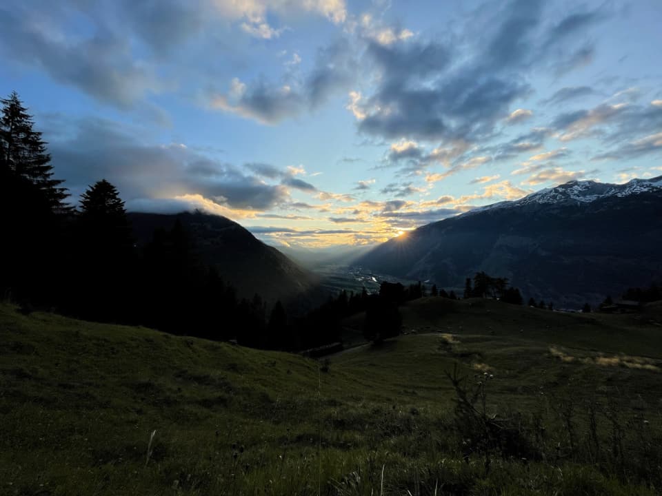 Sonnenuntergang über bergiger Landschaft mit Wiesen und Bäumen.