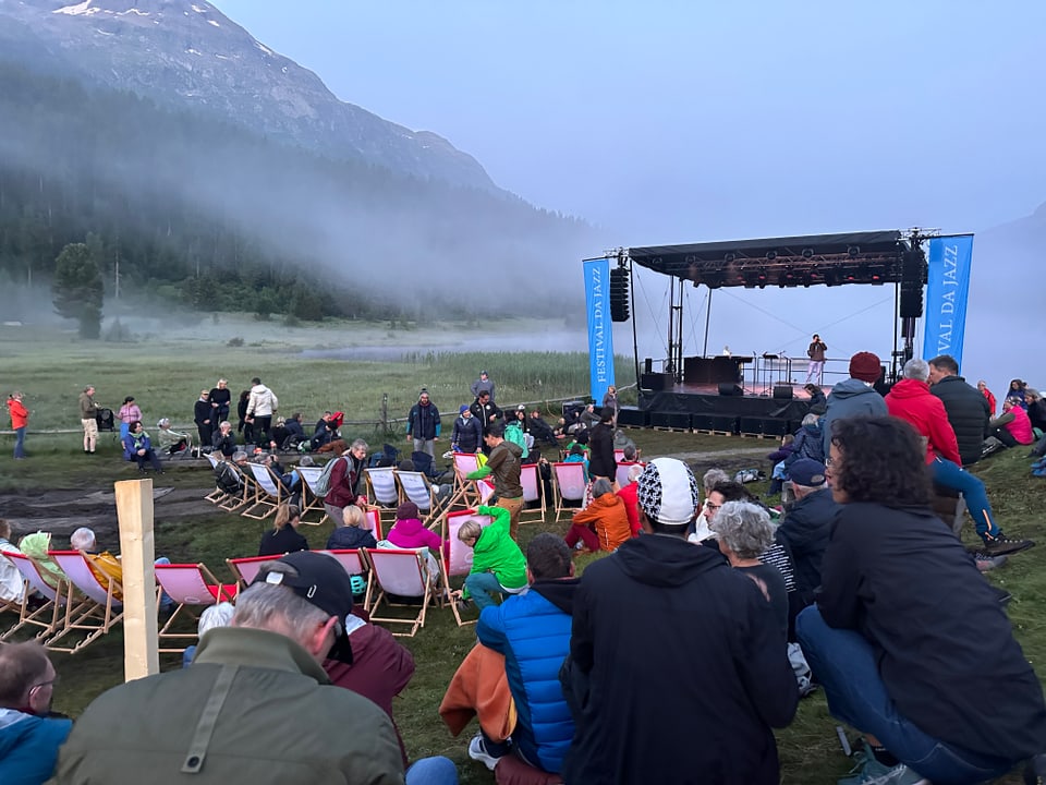 Outdoor-Konzert in bergiger Landschaft mit Publikum auf Stühlen.