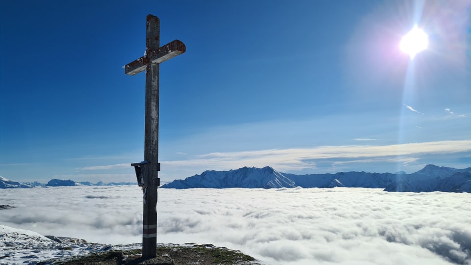 Ein Bergkreuz und ein Nebelmeer, dazu ein blauer Himmel.