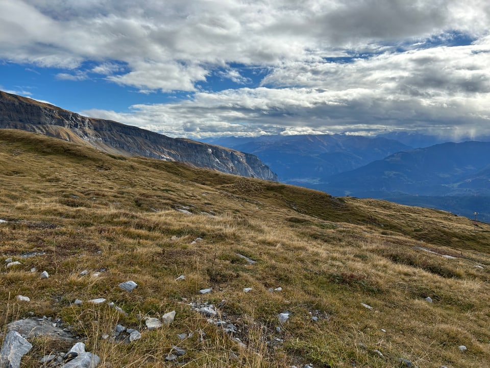 Grasbewachsener Hügel vor Berglandschaft unter wolkigem Himmel.