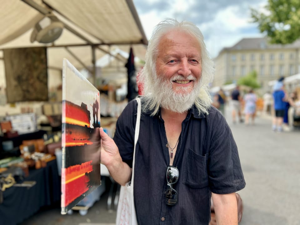 Älterer Mann mit langem weissem Bart hält ein Gemälde auf einem Markt.